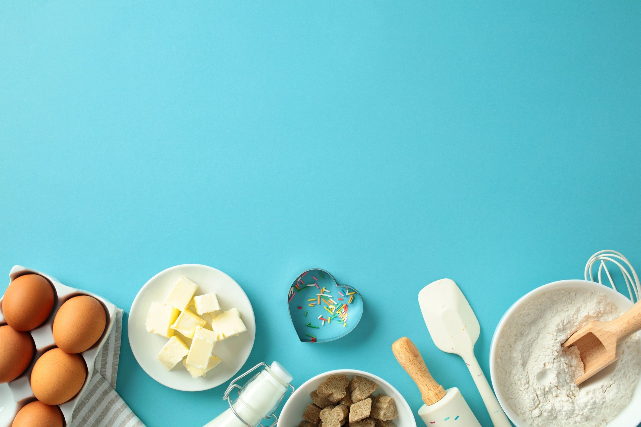 Ingredientes de repostería sobre fondo azul (huevos, mantequilla, leche, azúcar, harina y sprinkles)